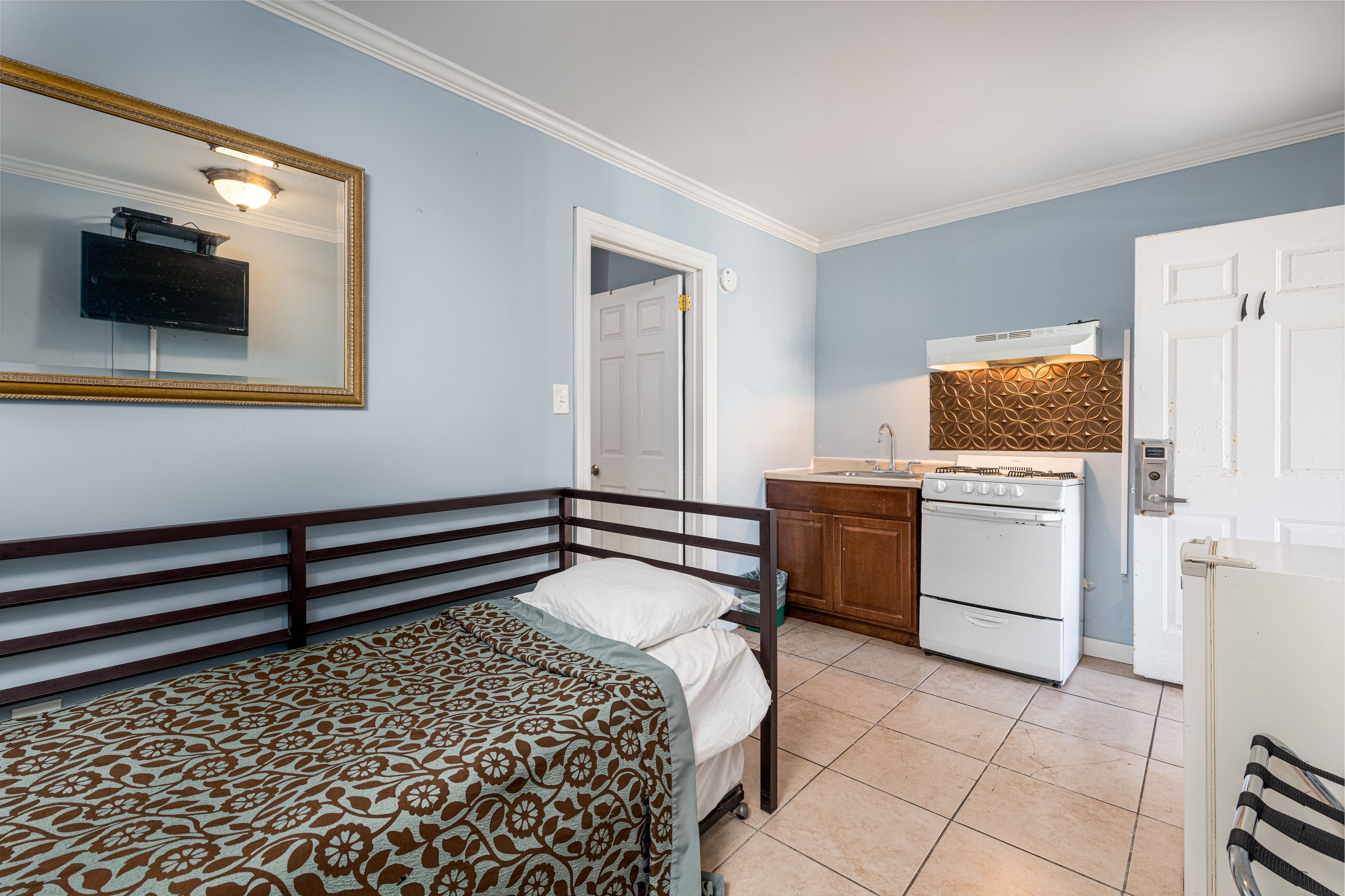 A bedroom with a bed, a dresser, and a mirror. There is a kitchen area with a stove and a refrigerator.
