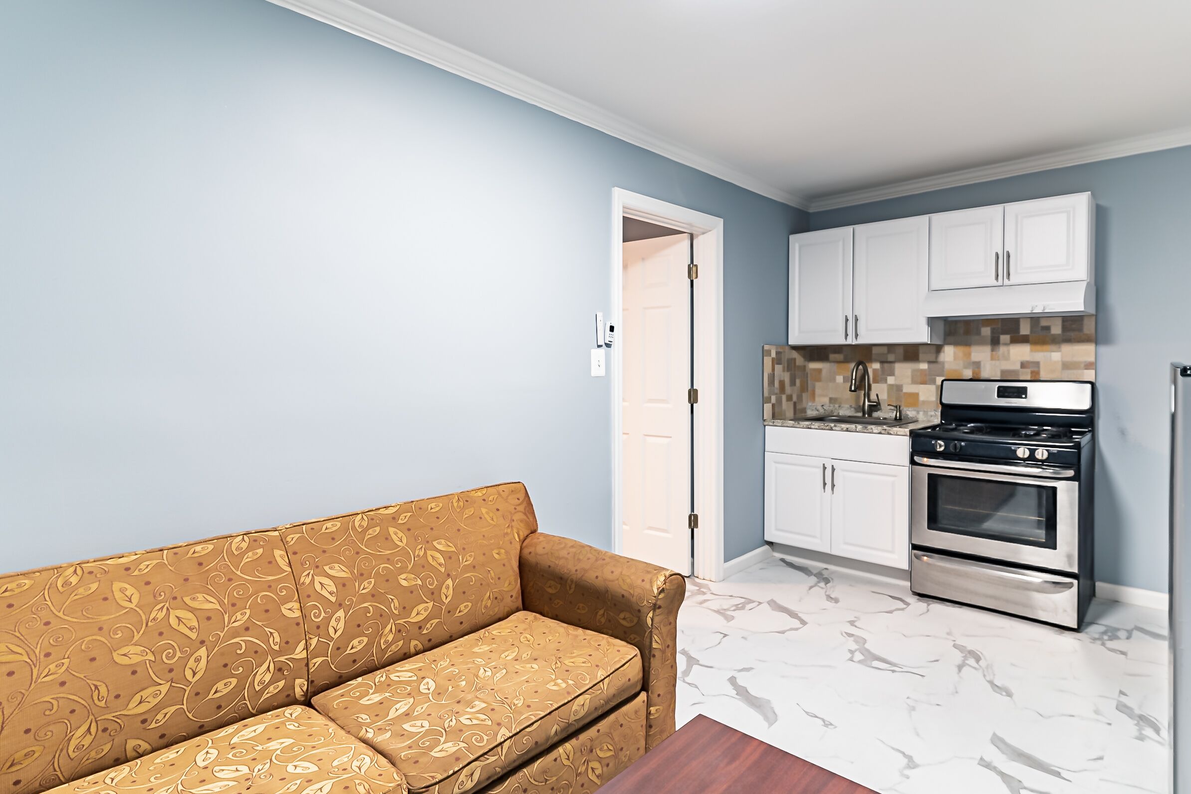 Una sala de estar con un sofá estampado, una cocina con armarios blancos y una estufa de acero inoxidable, y una puerta que da a otra habitación.