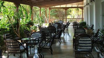 A covered outdoor seating area with wicker chairs and tables, surrounded by lush greenery and a wooden pergola.