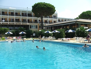 A swimming pool with lounge chairs and umbrellas, surrounded by a building with multiple balconies. - Irinna Hotel (Kefalonia)