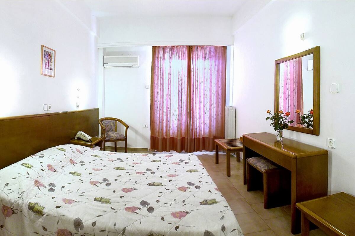 A hotel room with a bed, a desk, a chair, a bedside table with a vase of flowers, a mirror, and a window with curtains.