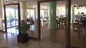 A spacious indoor area with a large glass door leading to an outdoor patio. Inside, there are several wooden chairs and tables arranged in a casual dining setting. A potted plant is placed near the entrance. - Irinna Hotel (Kefalonia)