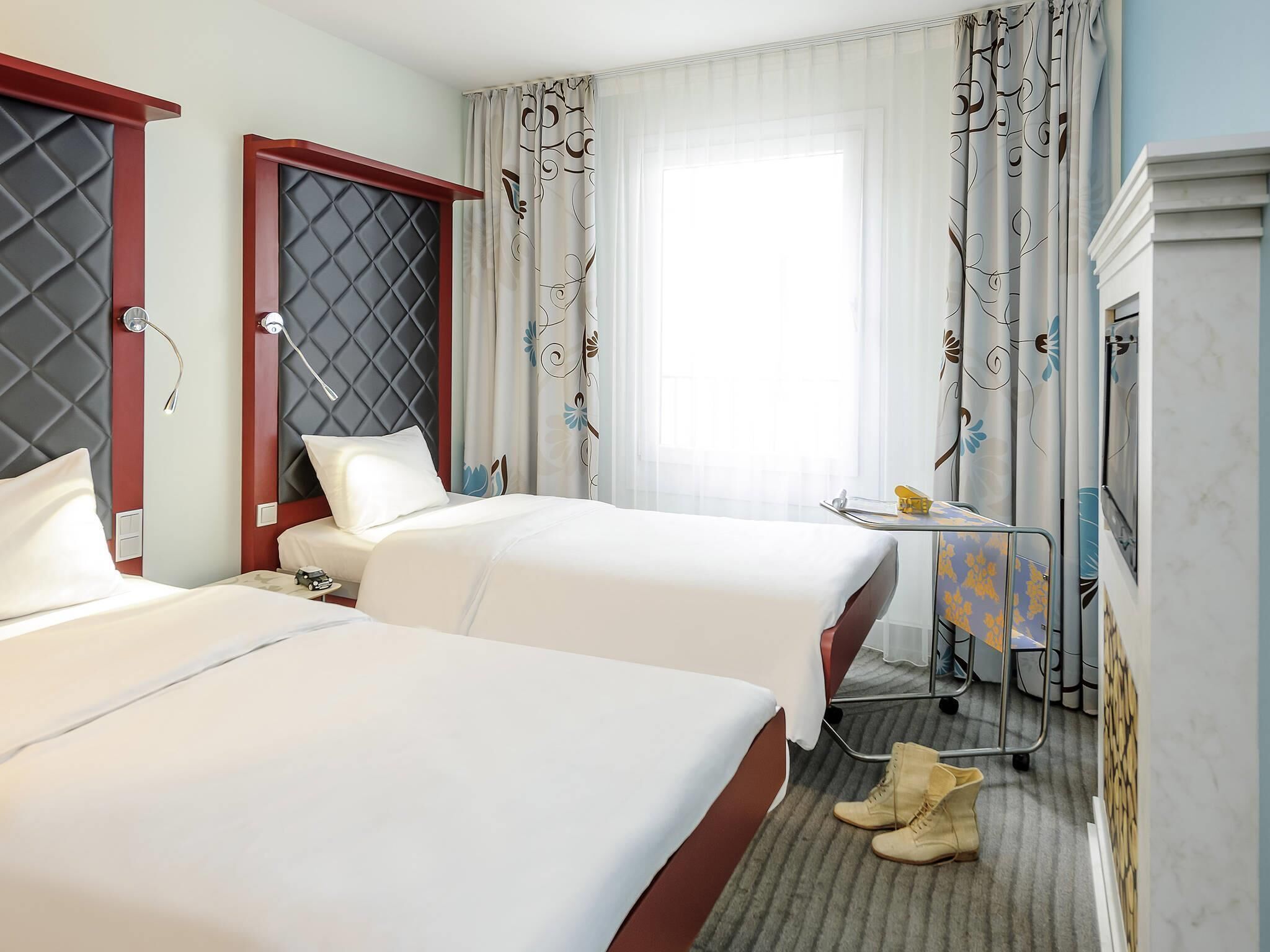 A hotel room with two beds, a small table with a chair, a pair of boots, and a window with curtains.