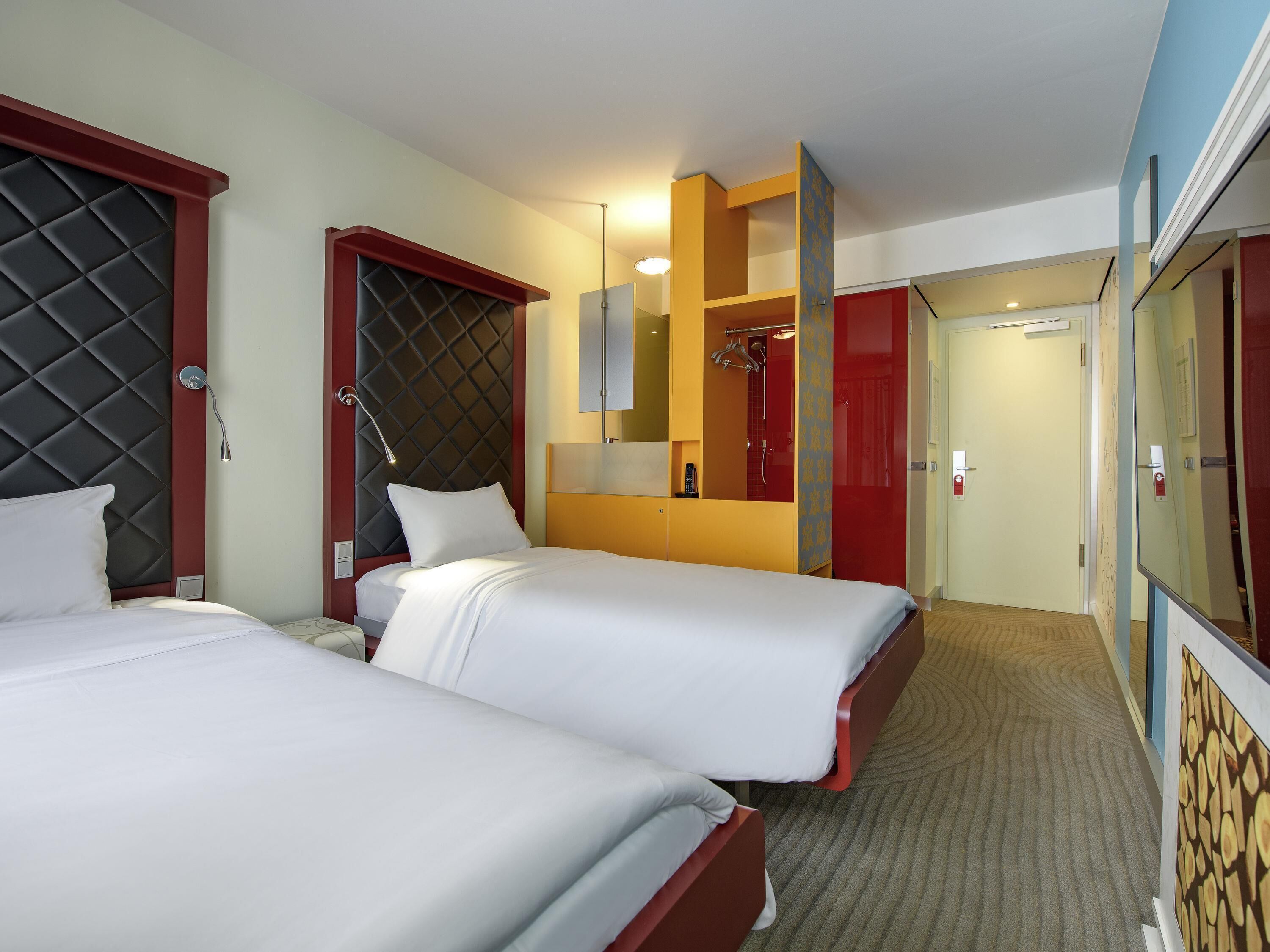 A hotel room with two beds, a red wardrobe, a mirror, and a shower area.