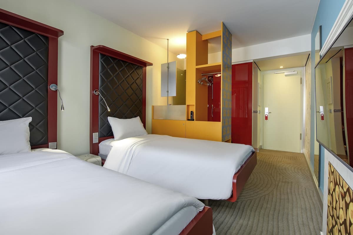 A hotel room with two beds, a red wardrobe, a mirror, and a shower area.