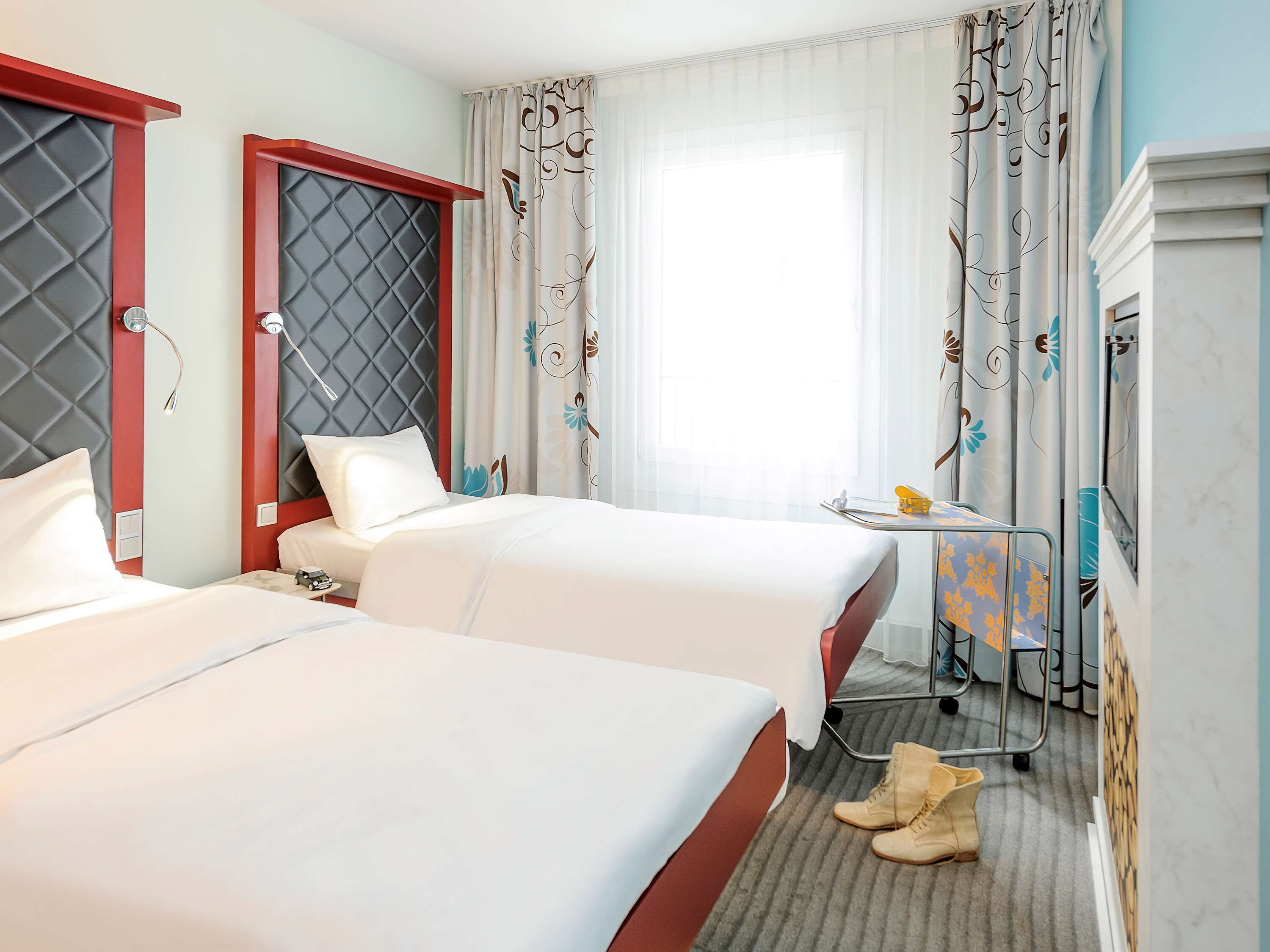 A hotel room with two beds, a small table with a lamp, and a window with curtains.