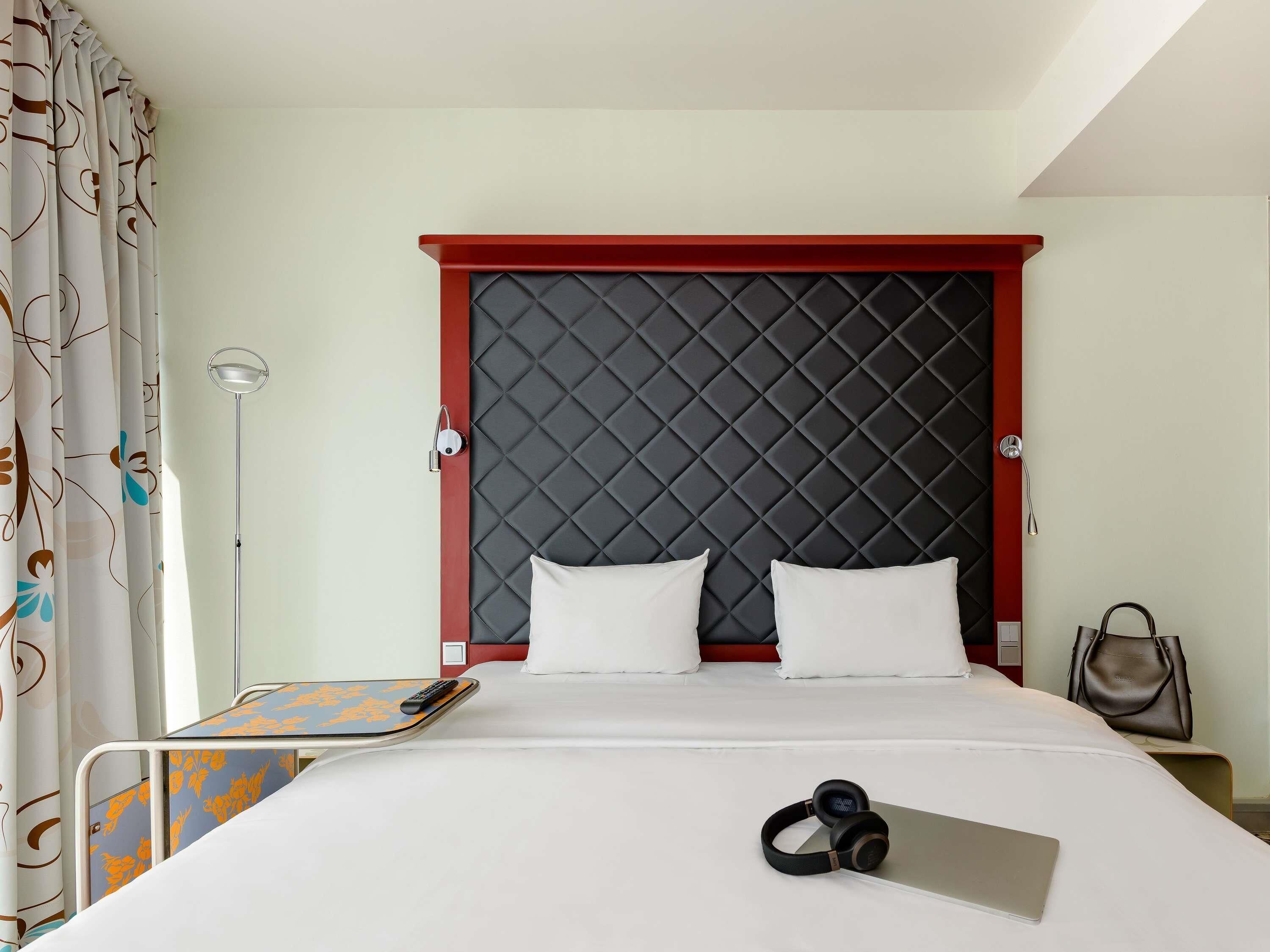 Una habitación de hotel con una cama, una mesita de noche, una lámpara, una maleta y auriculares sobre la cama.