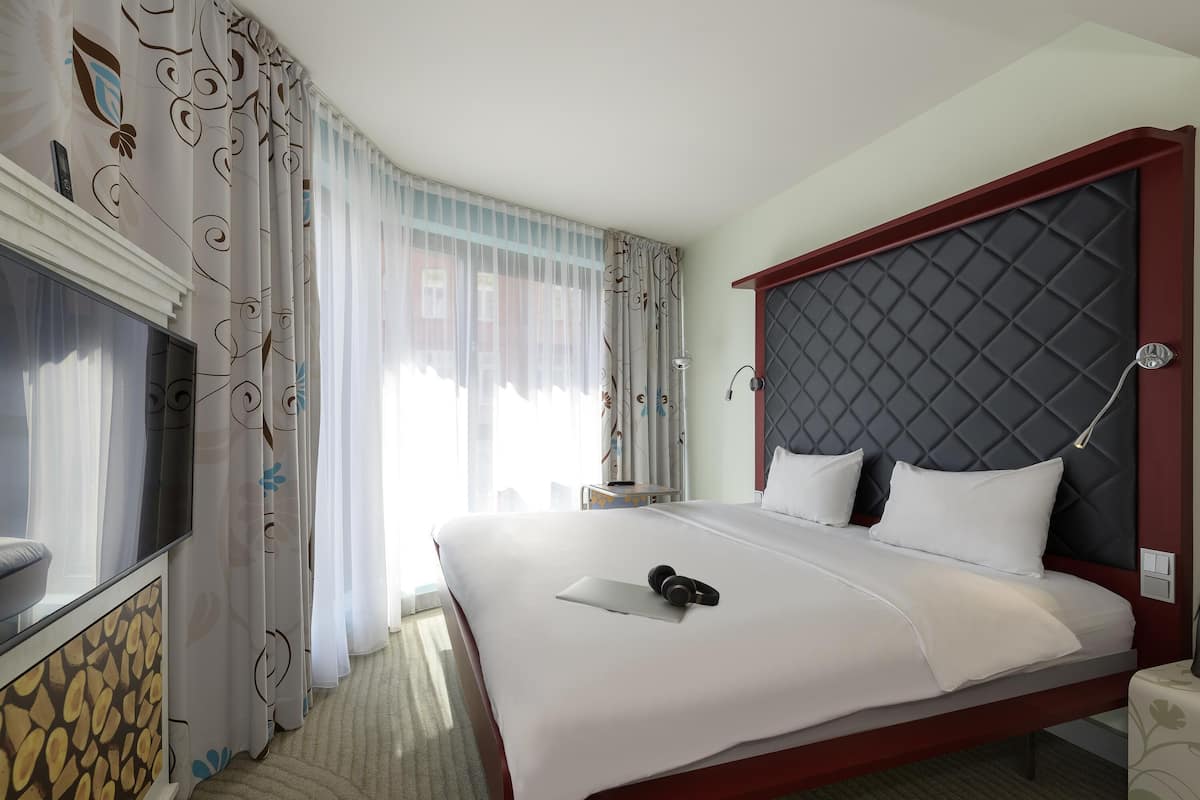 A hotel room with a large bed, a TV, a bedside table, and a window with curtains.