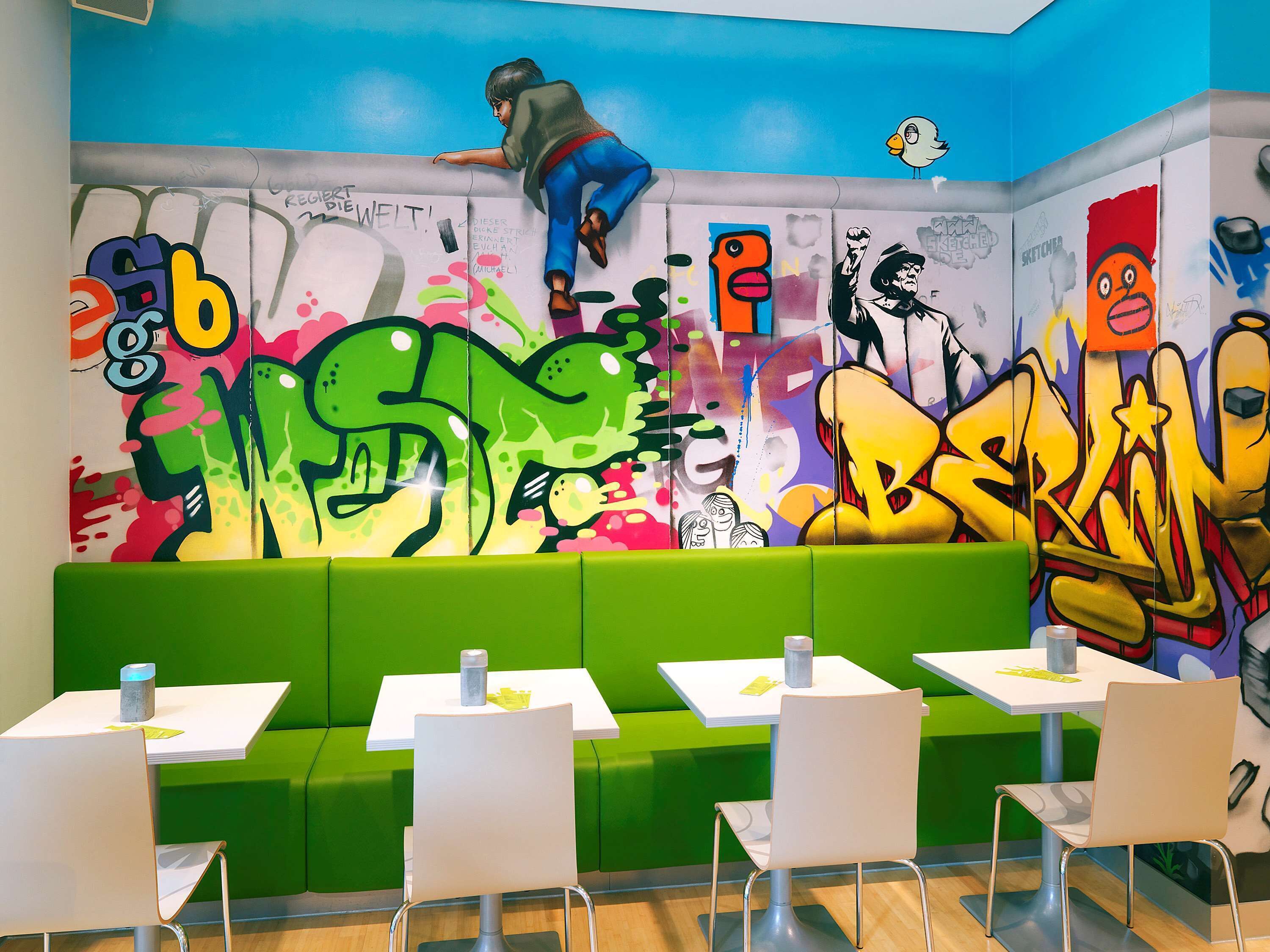 A vibrant graffiti wall with colorful artwork and a person climbing it. There are green seating areas and white tables with chairs.