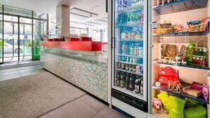 Un vestíbulo de hotel con una recepción, un refrigerador de bebidas repleto y estantes con aperitivos y artículos de aseo.