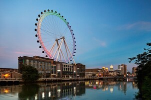A large Ferris wheel with a reflection in the water, surrounded by buildings and a bridge.