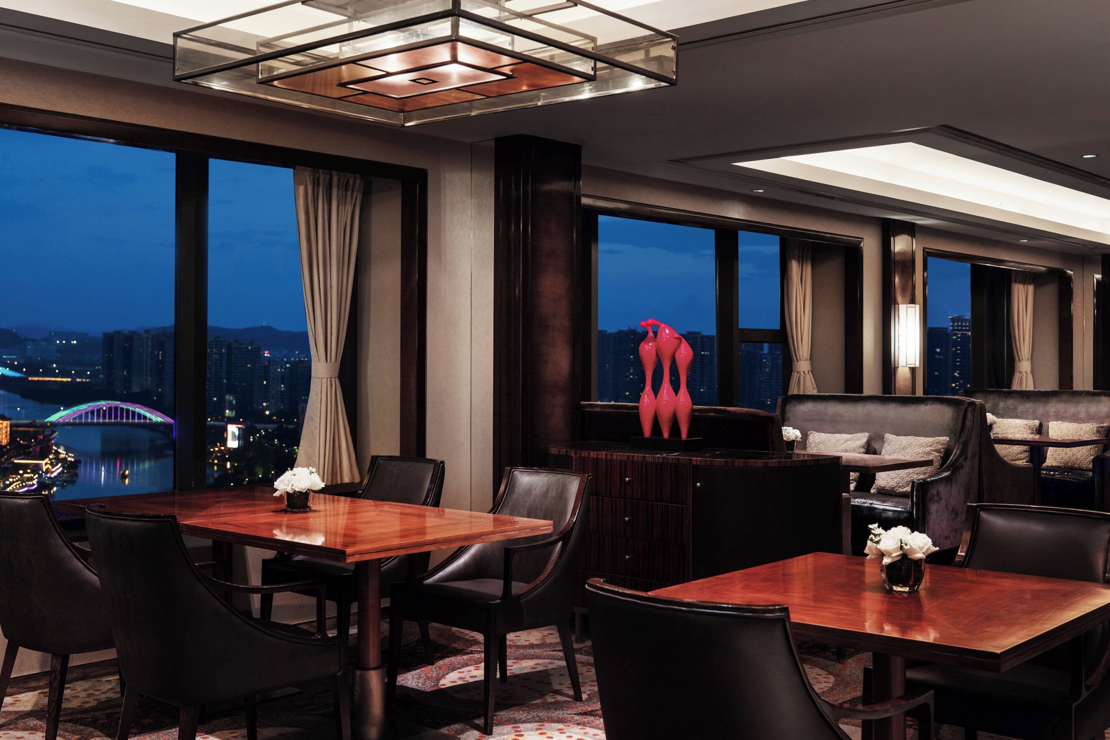 A modern hotel room with a dining area, a sofa set, and large windows offering a city view.