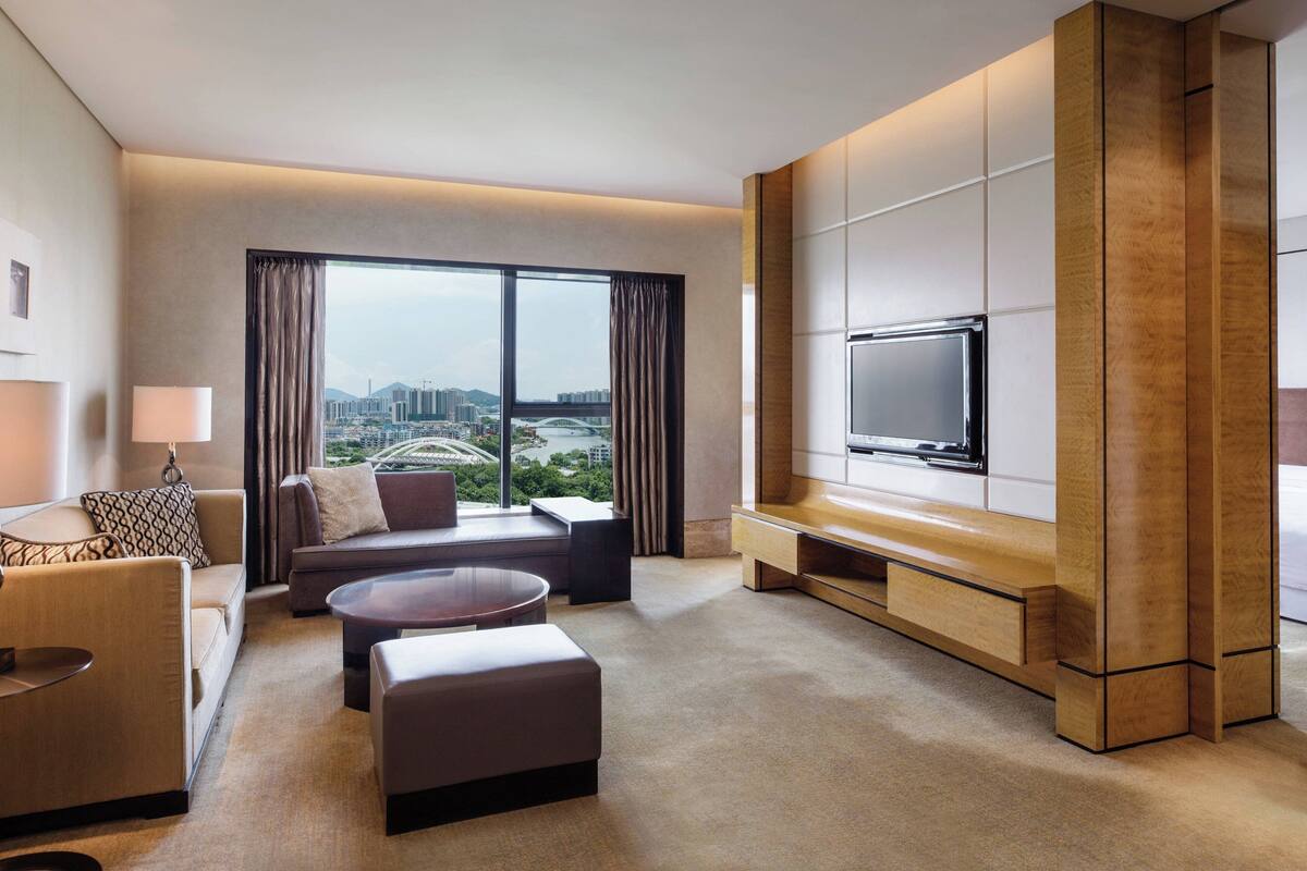 Habitación de hotel moderna con un ventanal grande que ofrece vistas a la ciudad, un televisor de pantalla plana montado en un panel de madera, un sofá, una mesa de centro y una mesita auxiliar.