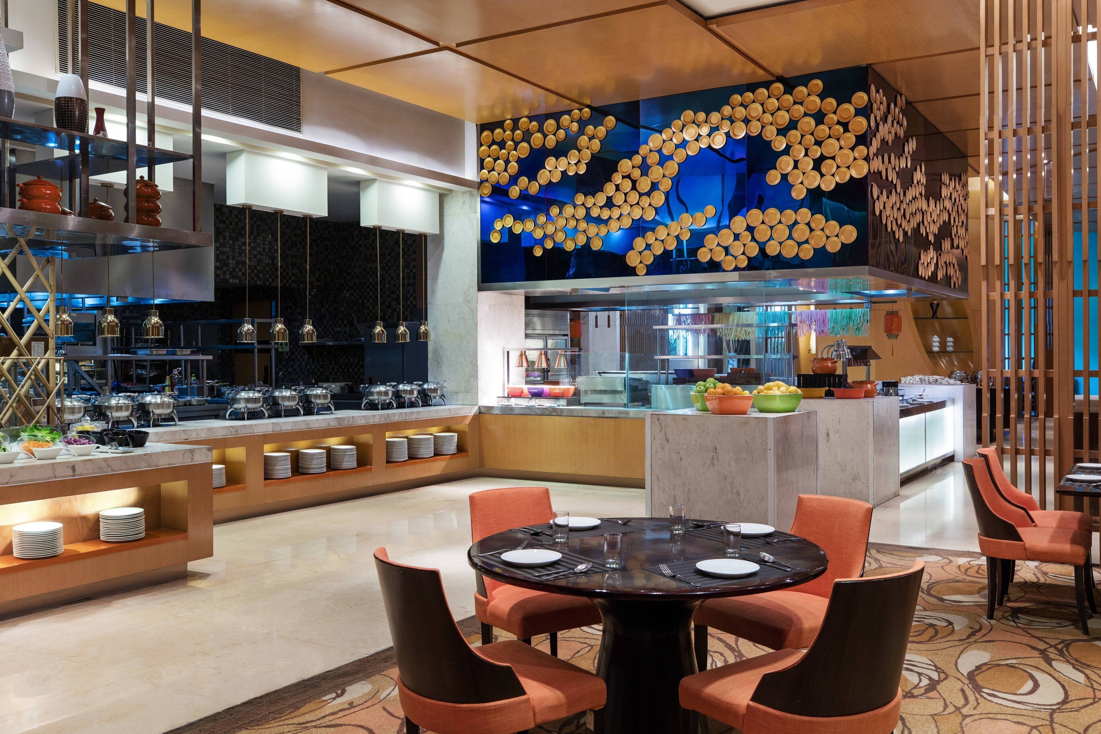 A modern hotel lobby with a central dining area, a kitchen with a buffet, and a decorative ceiling.