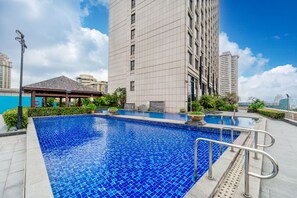 Una piscina en la azotea con superficie de azulejos azules, rodeada por un edificio alto, un área de estar techada y algo de vegetación.