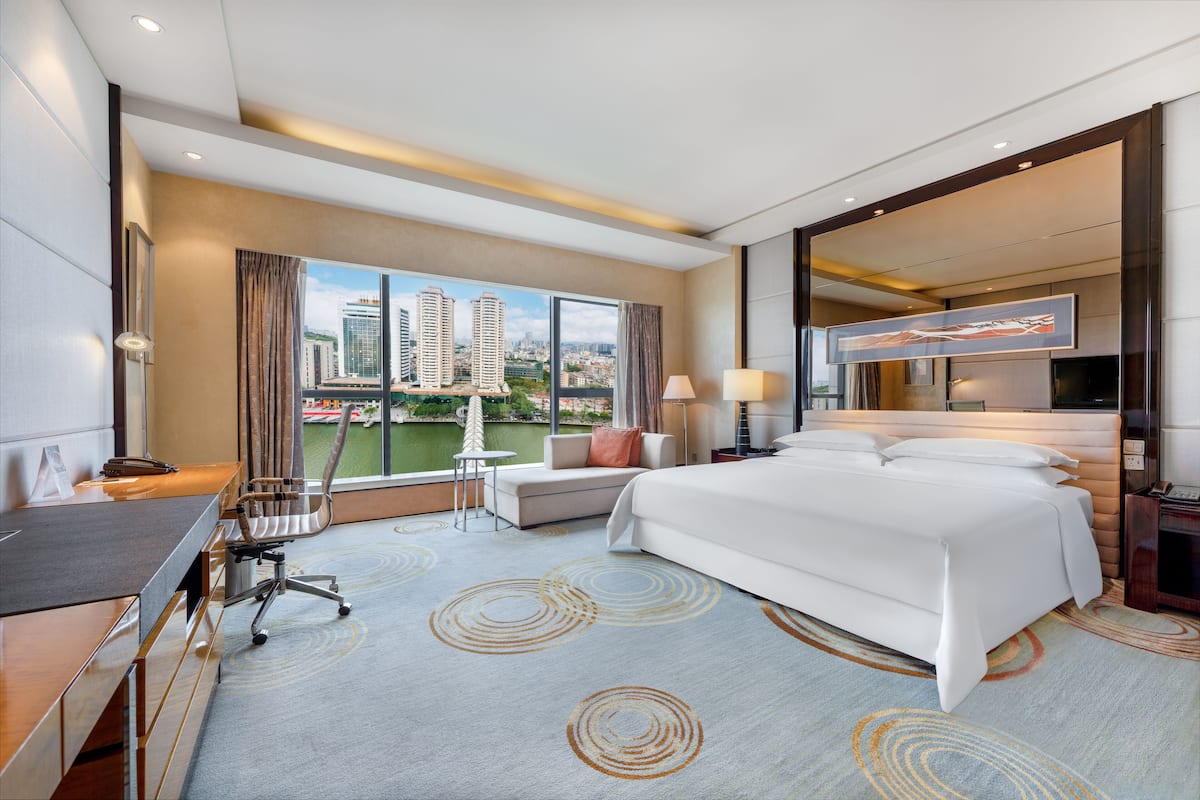 Una habitación de hotel moderna con una cama grande, un escritorio, una silla y vistas de la ciudad a través de amplias ventanas.