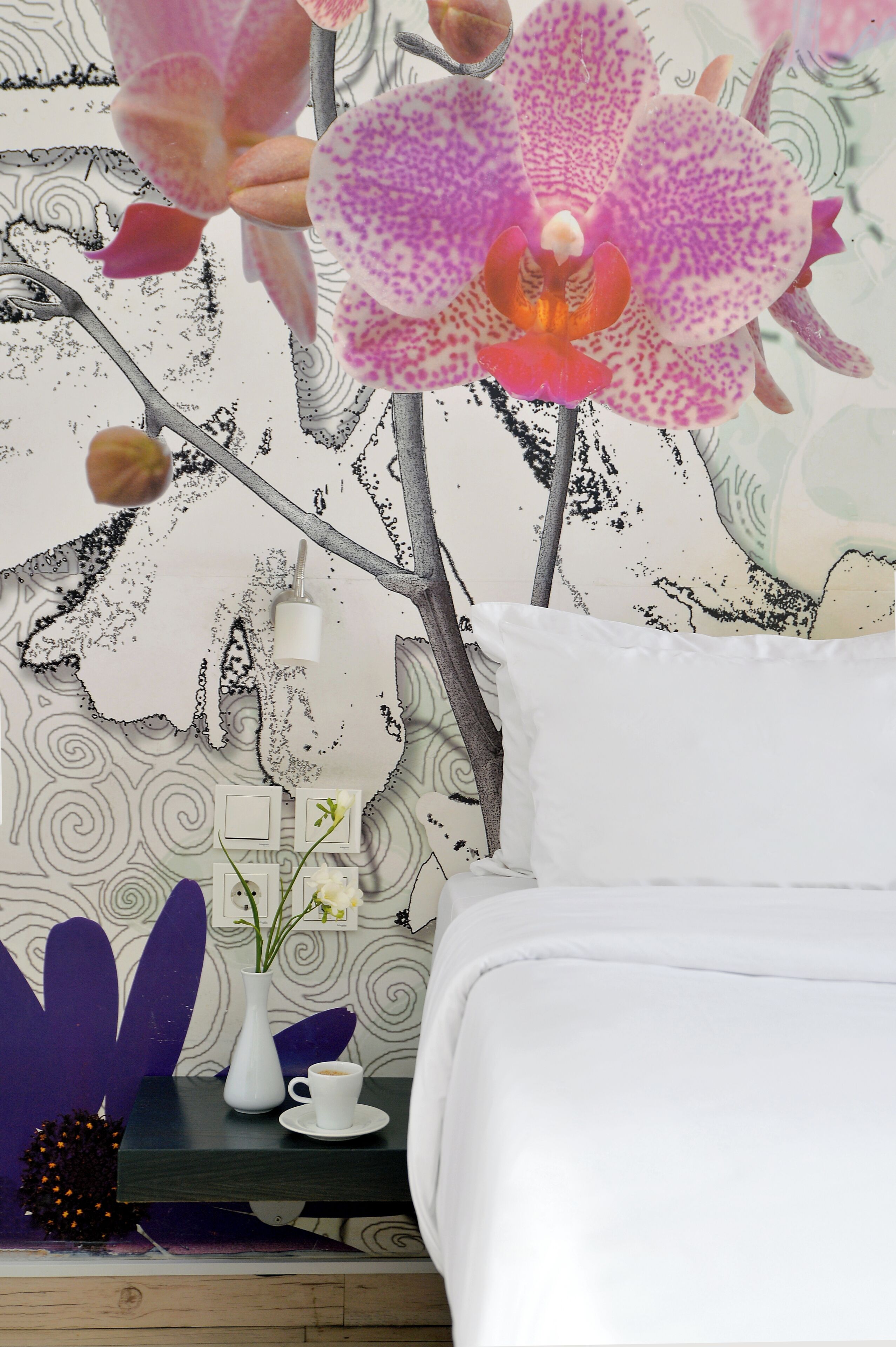 A bedroom with a bed, a nightstand with a vase and a cup, and a wall with a floral pattern.