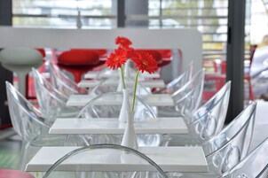 Un comedor moderno con mesas y sillas de vidrio transparente, un jarrón blanco con flores rojas, y una silla roja al fondo.