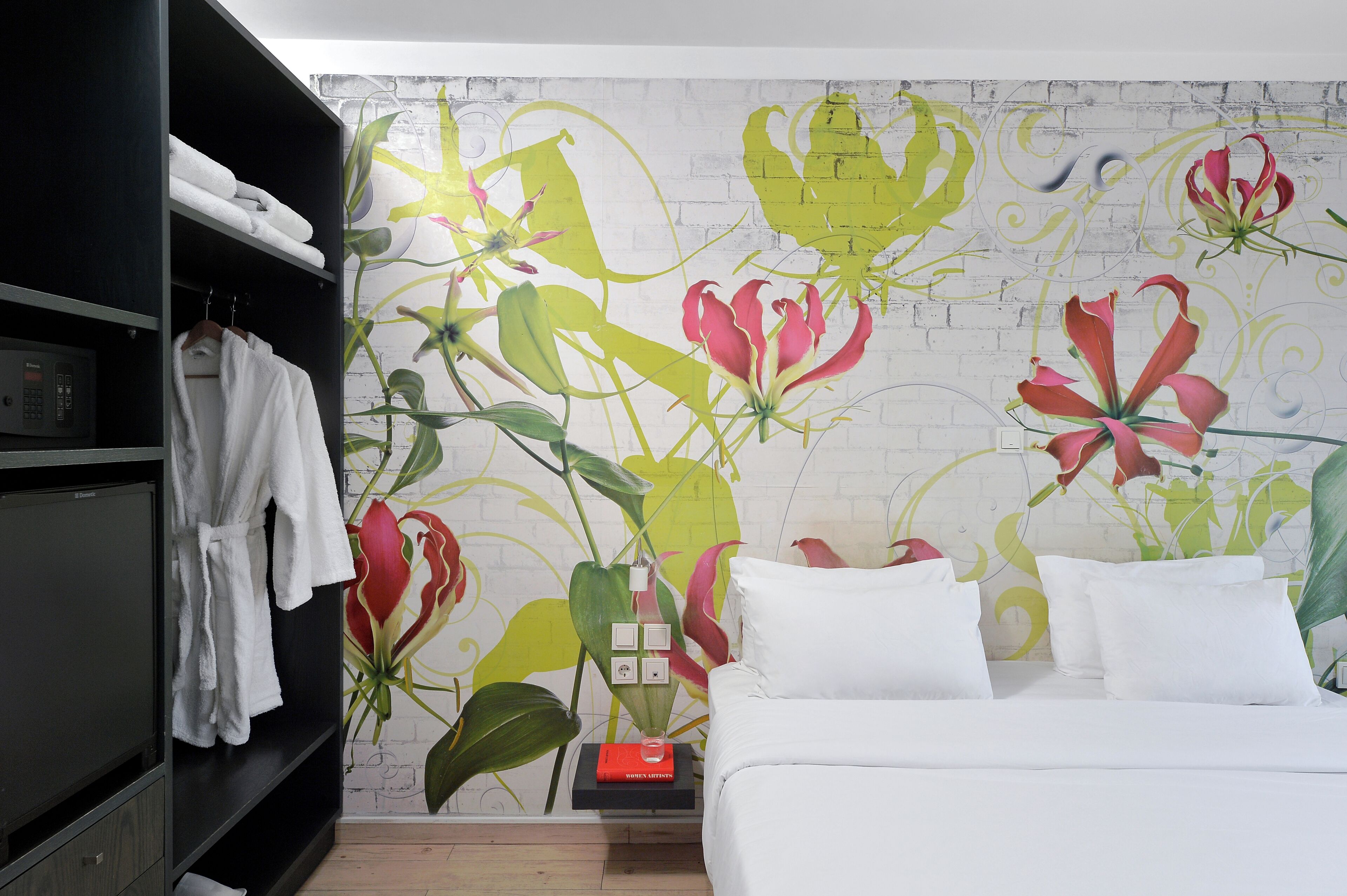 Quarto de hotel com cama branca, guarda-roupa preto e parede com um mural floral.