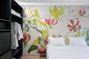 Quarto de hotel com cama branca, guarda-roupa preto e parede com um mural floral.