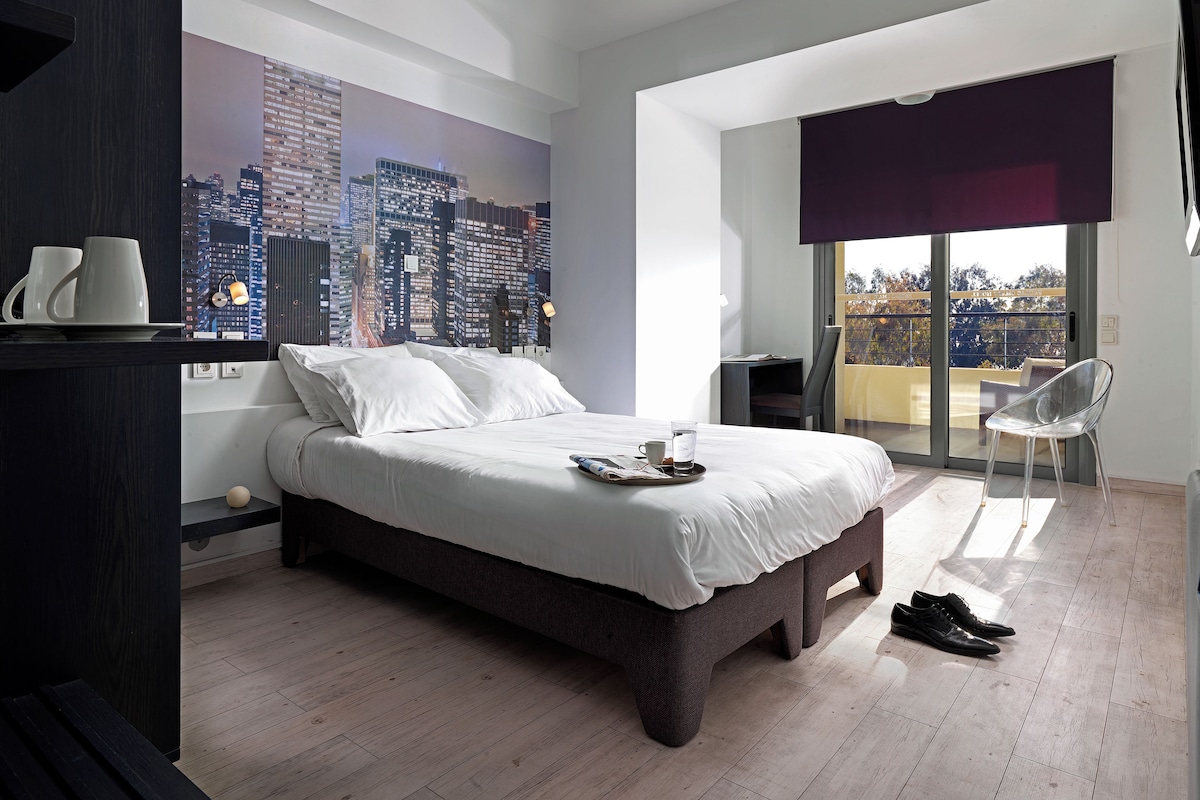 A modern hotel room with a large bed, a cityscape mural, a balcony with a view, and a dining area with a table and chairs.