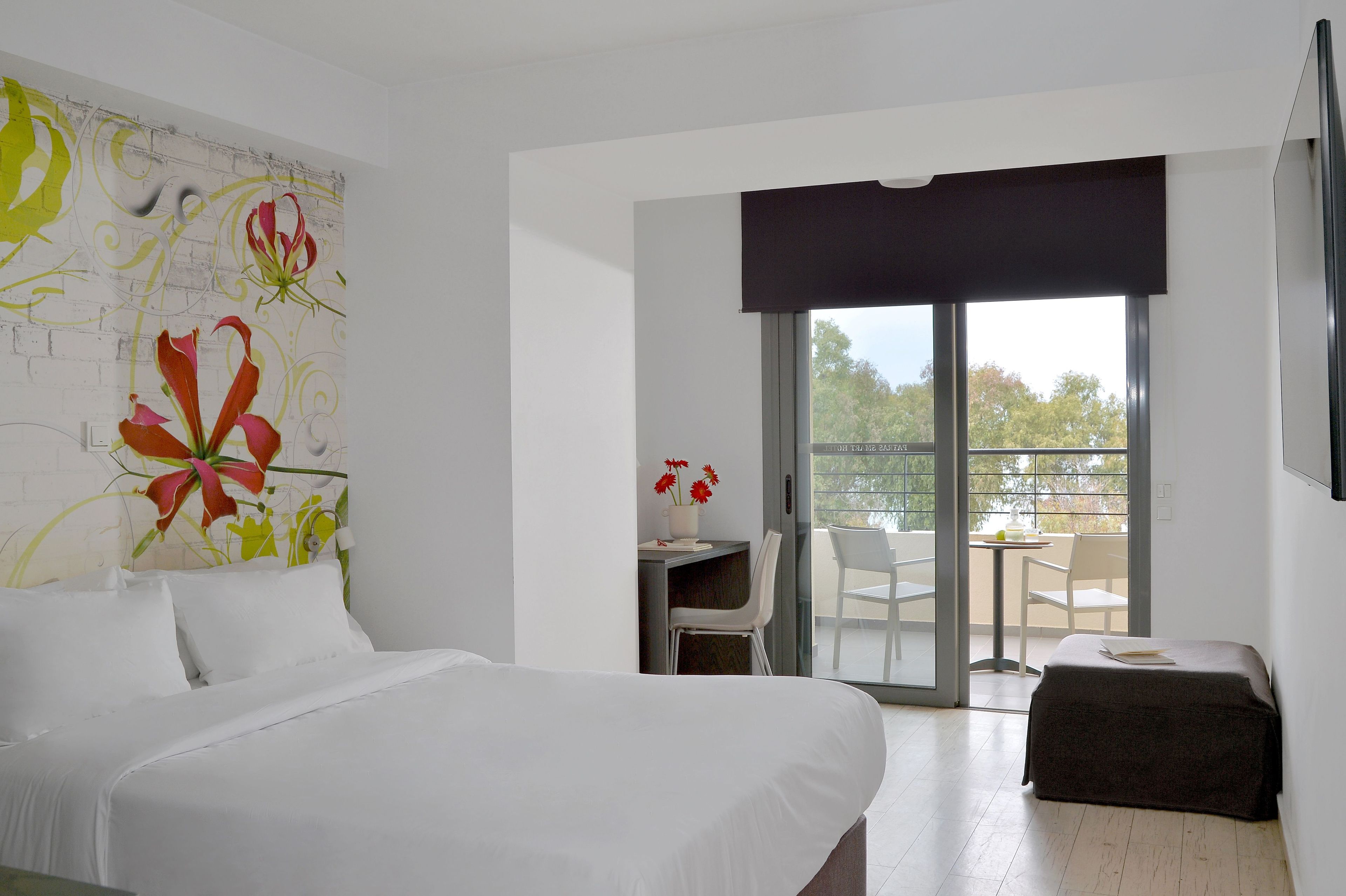 Habitación de hotel con cama, escritorio con silla, balcón con mesa y sillas, y una ventana grande.
