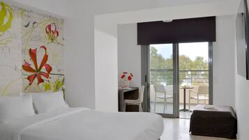 A hotel room with a bed, a desk with a chair, a balcony with a table and chairs, and a large window.