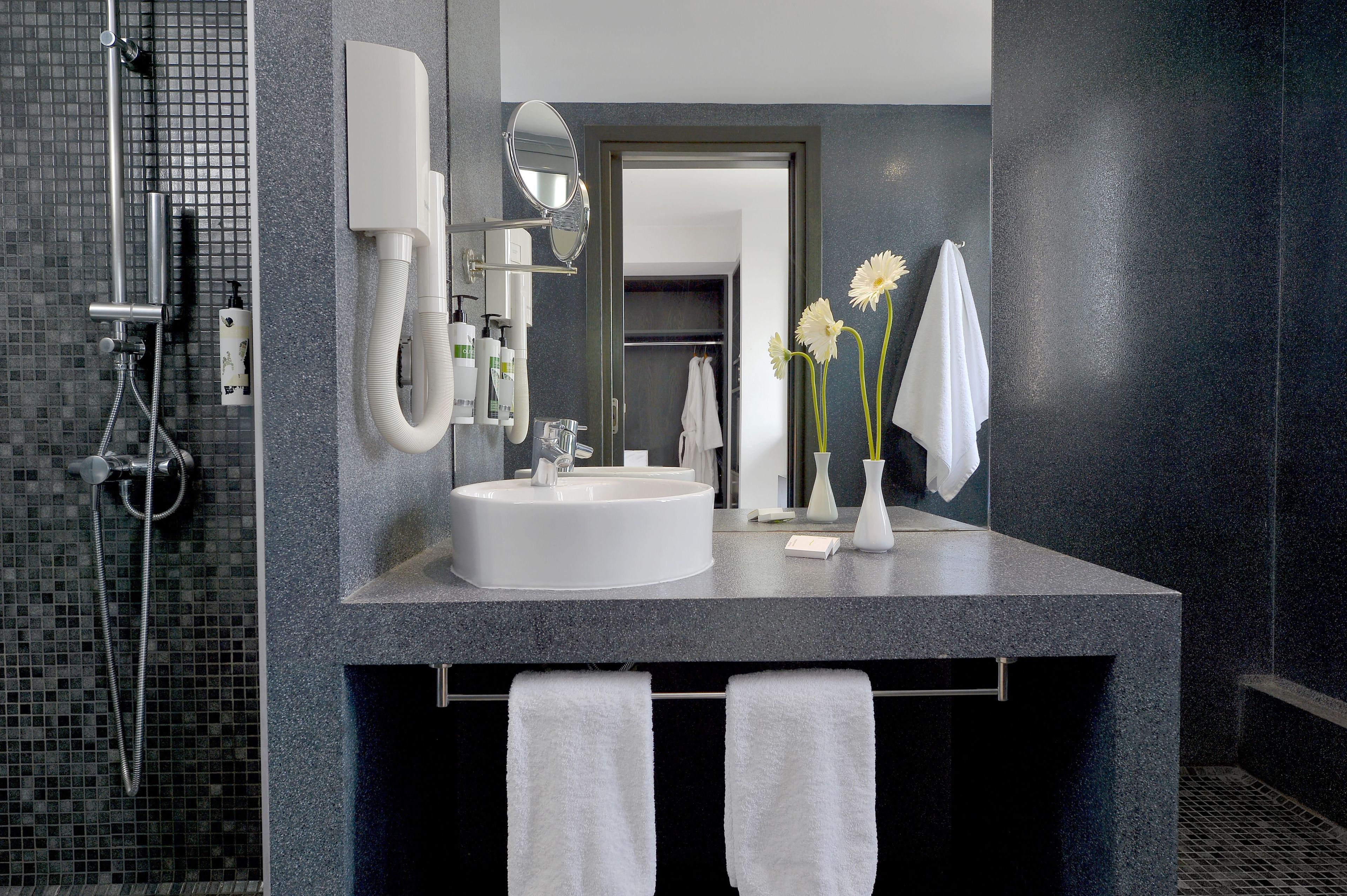 Een moderne badkamer met een grote spiegel, een wastafel en een douche. Handdoeken hangen aan een rek en op het aanrecht staat een vaas met bloemen.