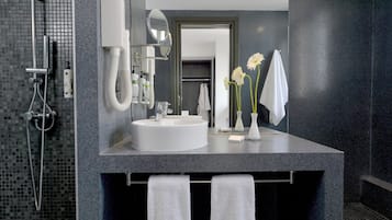 Een moderne badkamer met een grote spiegel, een wastafel en een douche. Handdoeken hangen aan een rek en op het aanrecht staat een vaas met bloemen.