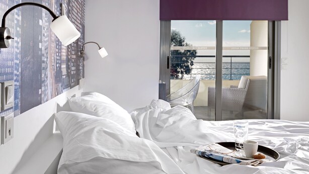 Habitación de hotel con cama, ropa blanca, mesita de noche, lámpara, bandeja con vaso de agua, taza y un muffin.