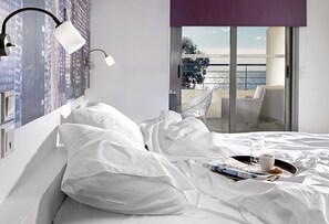 Habitación de hotel con cama, ropa blanca, mesita de noche, lámpara, bandeja con vaso de agua, taza y un muffin.