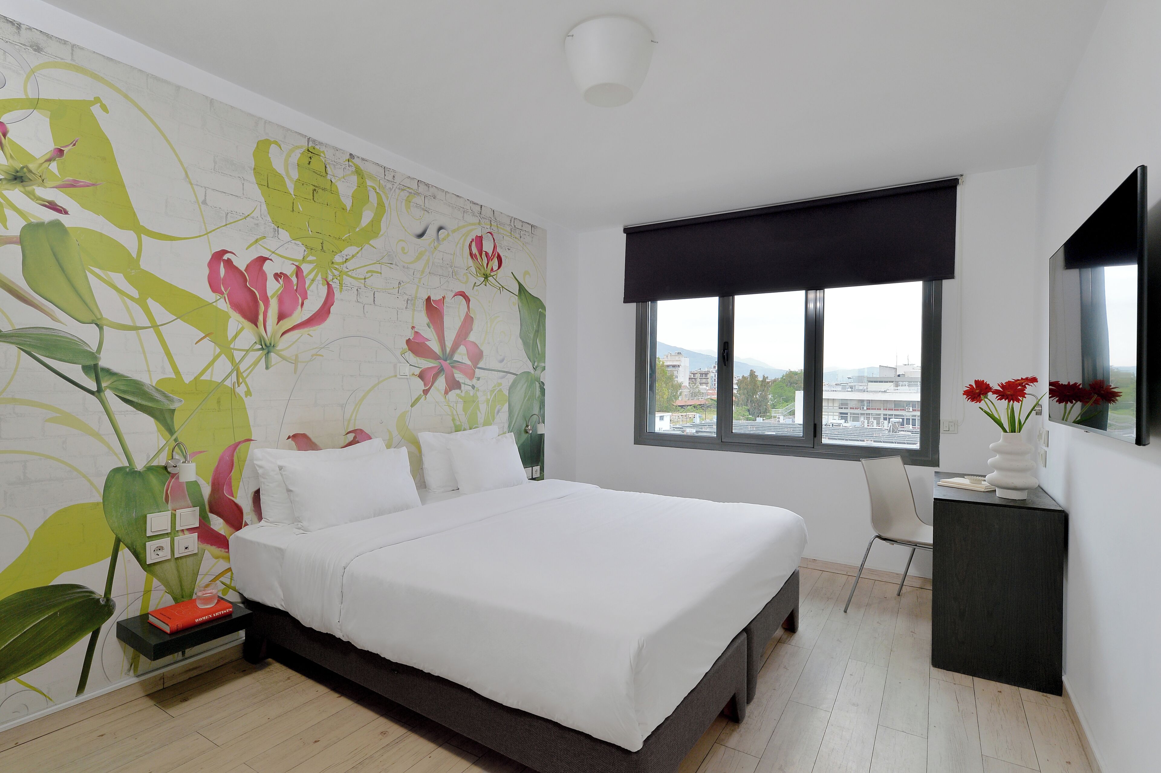 Un dormitorio con una cama grande, una mesita de noche, un escritorio, una silla, un ventanal con vistas y una pared con un mural floral.