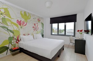 Un dormitorio con una cama grande, una mesita de noche, un escritorio, una silla, un ventanal con vistas y una pared con un mural floral.