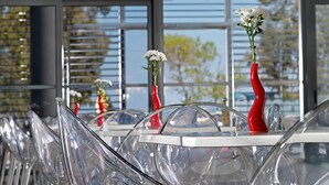 A modern dining area with clear glass chairs and tables, red vases with white flowers, and large windows offering a view of greenery. - Airotel Patras Smart (Patras)
