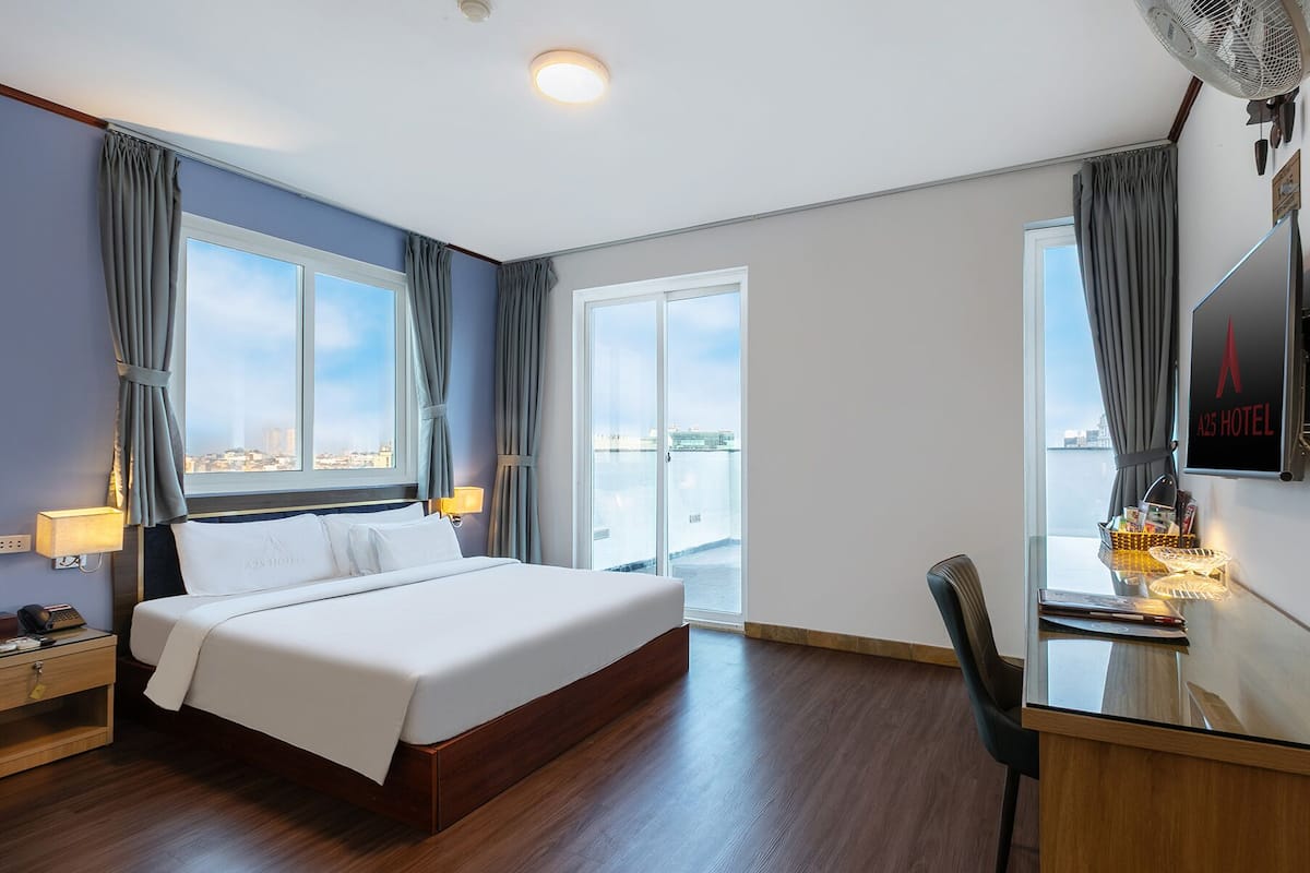 A hotel room with a bed, a desk, a chair, a TV, and a view of the city through large windows.