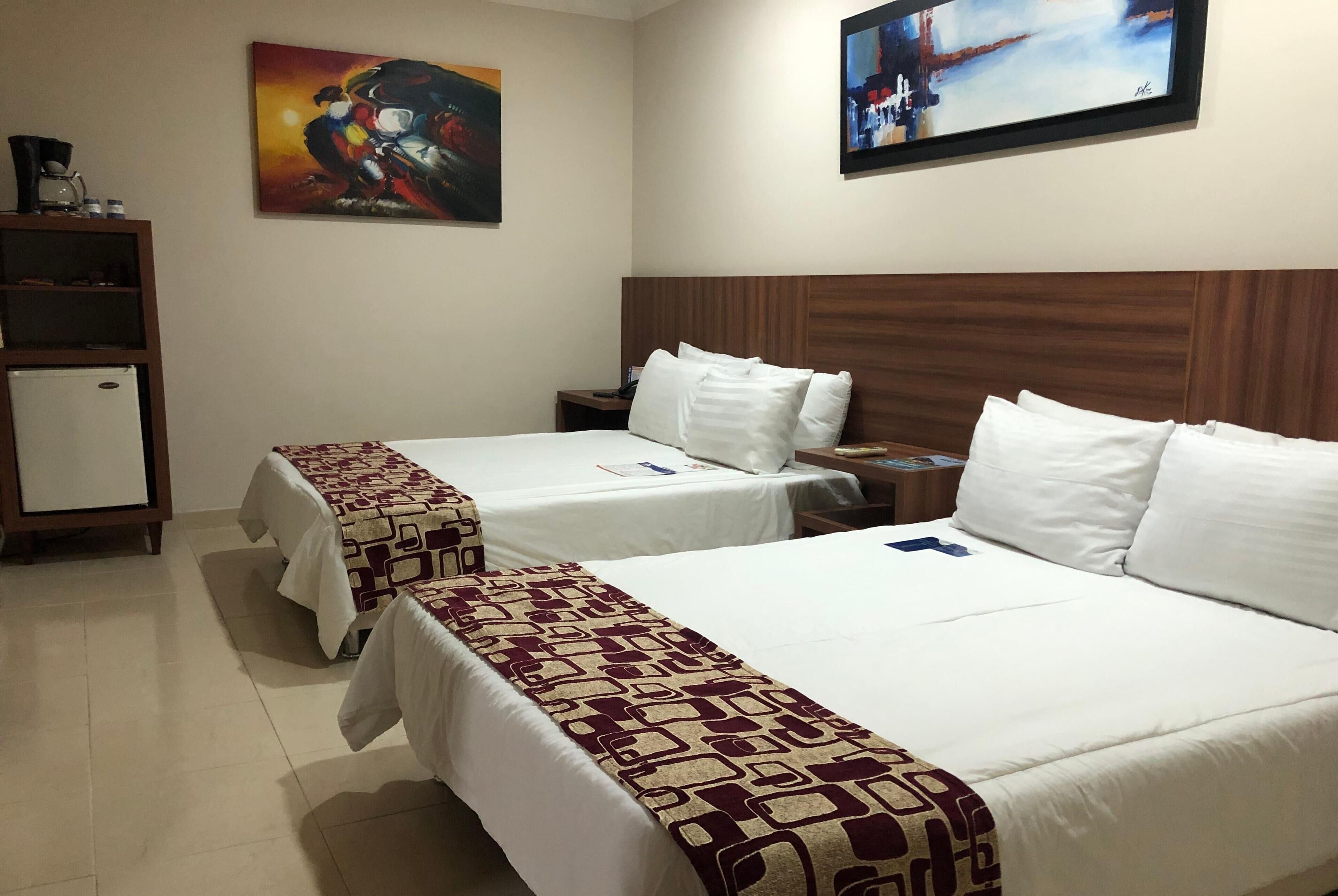 Habitación de hotel con dos camas, cabecera de madera, un mueble pequeño y obras de arte en las paredes.