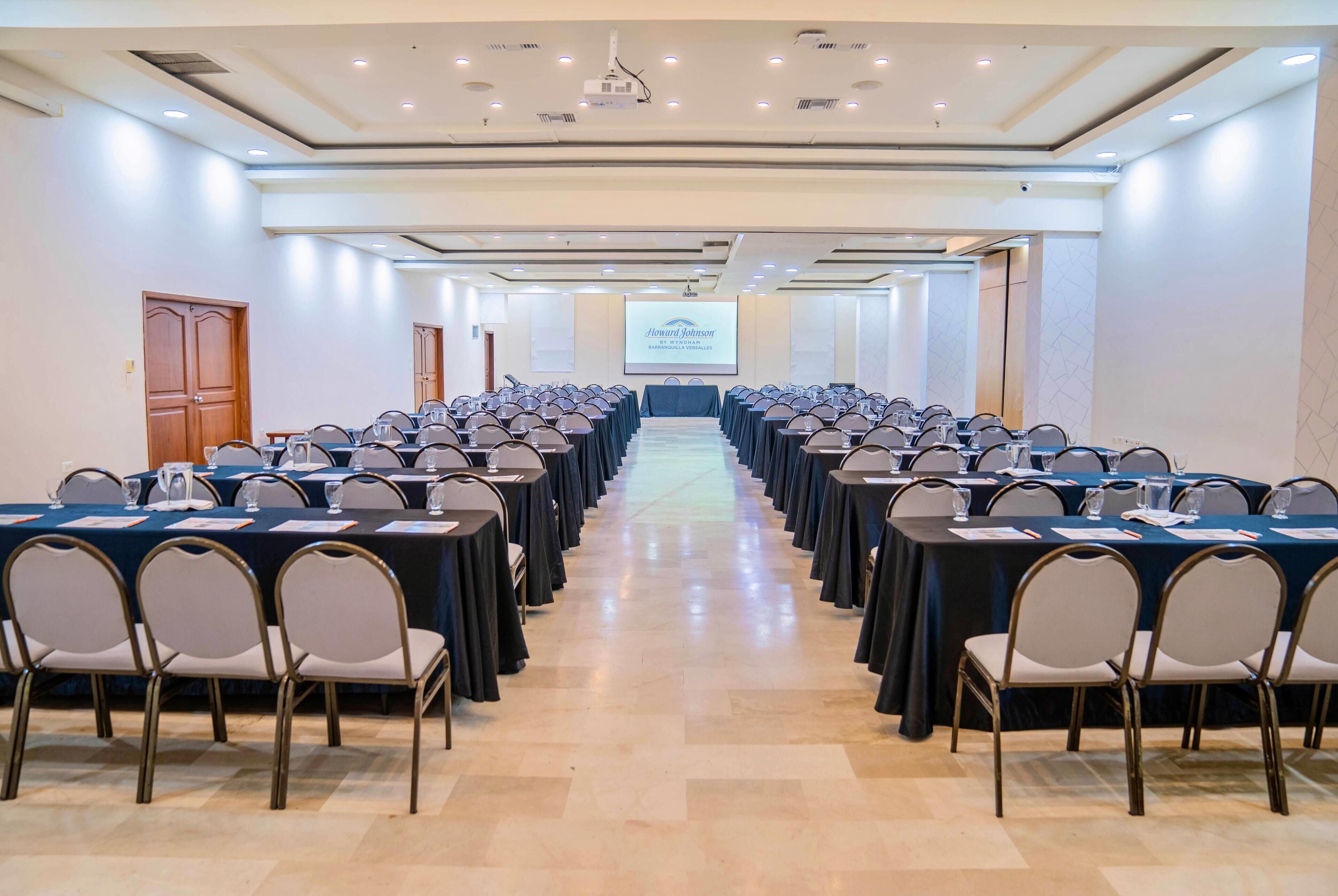 Une salle de conférence avec des rangées de tables et de chaises, un écran de projection et une porte sur le côté gauche.