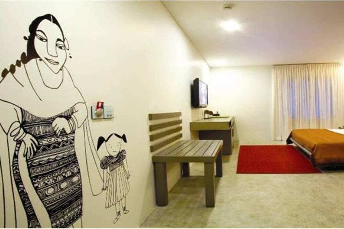 A hotel room with a bed, a desk, a chair, a television, and a wall with a drawing.