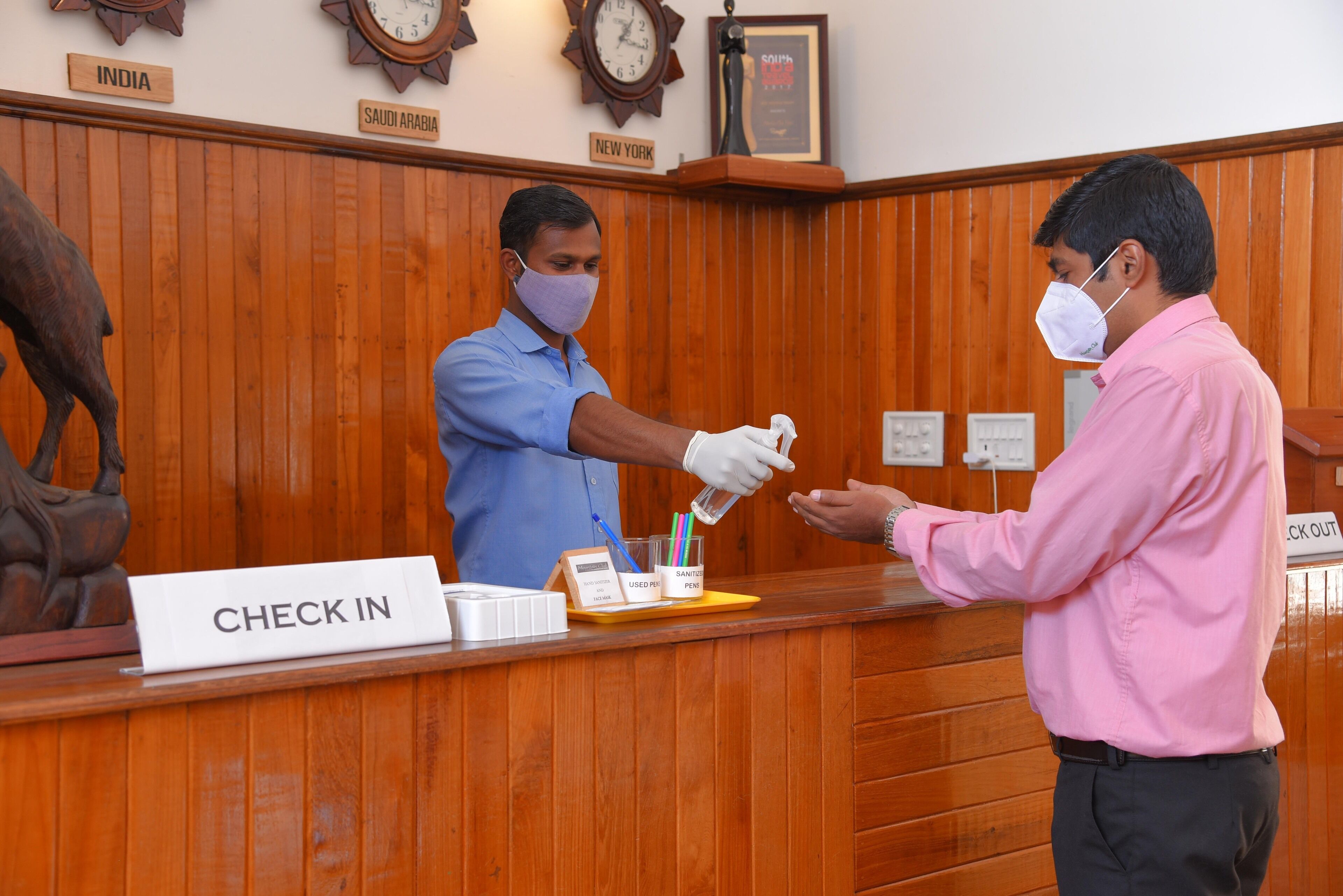 A reception desk with a "CHECK IN" sign, a clock, and wall-mounted signs indicating "INDIA," "SAUDI ARABIA," and "NEW YORK."