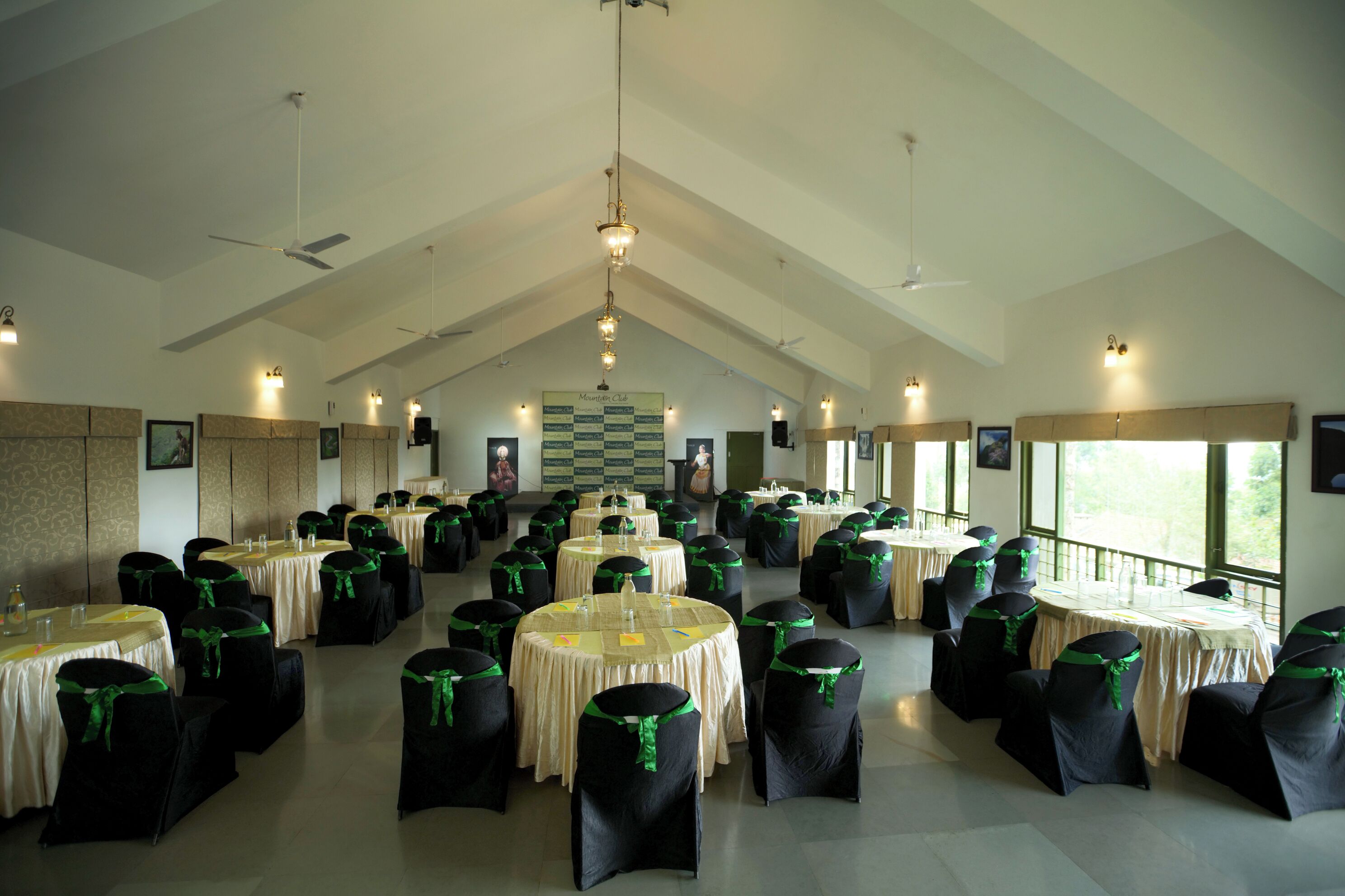Un salón amplio con varias mesas redondas, cada una cubierta con un mantel blanco y sillas negras adornadas con cintas verdes.