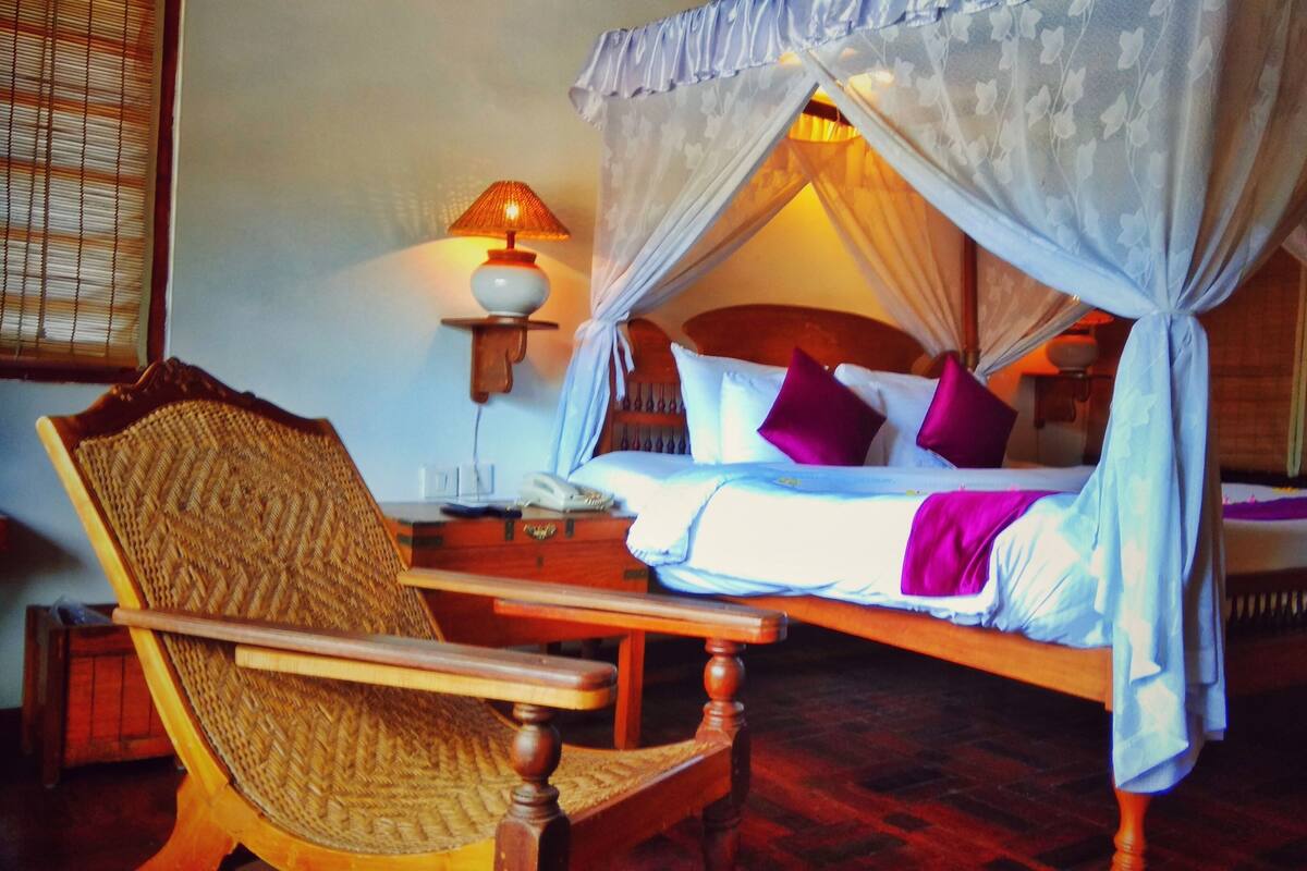 A room with a four-poster bed, a bedside table with a lamp, and a wicker chair.