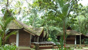 A traditional wooden structure with a tiled roof, surrounded by lush greenery and palm trees. - Punnamada Resorts (Ambalapuzha)