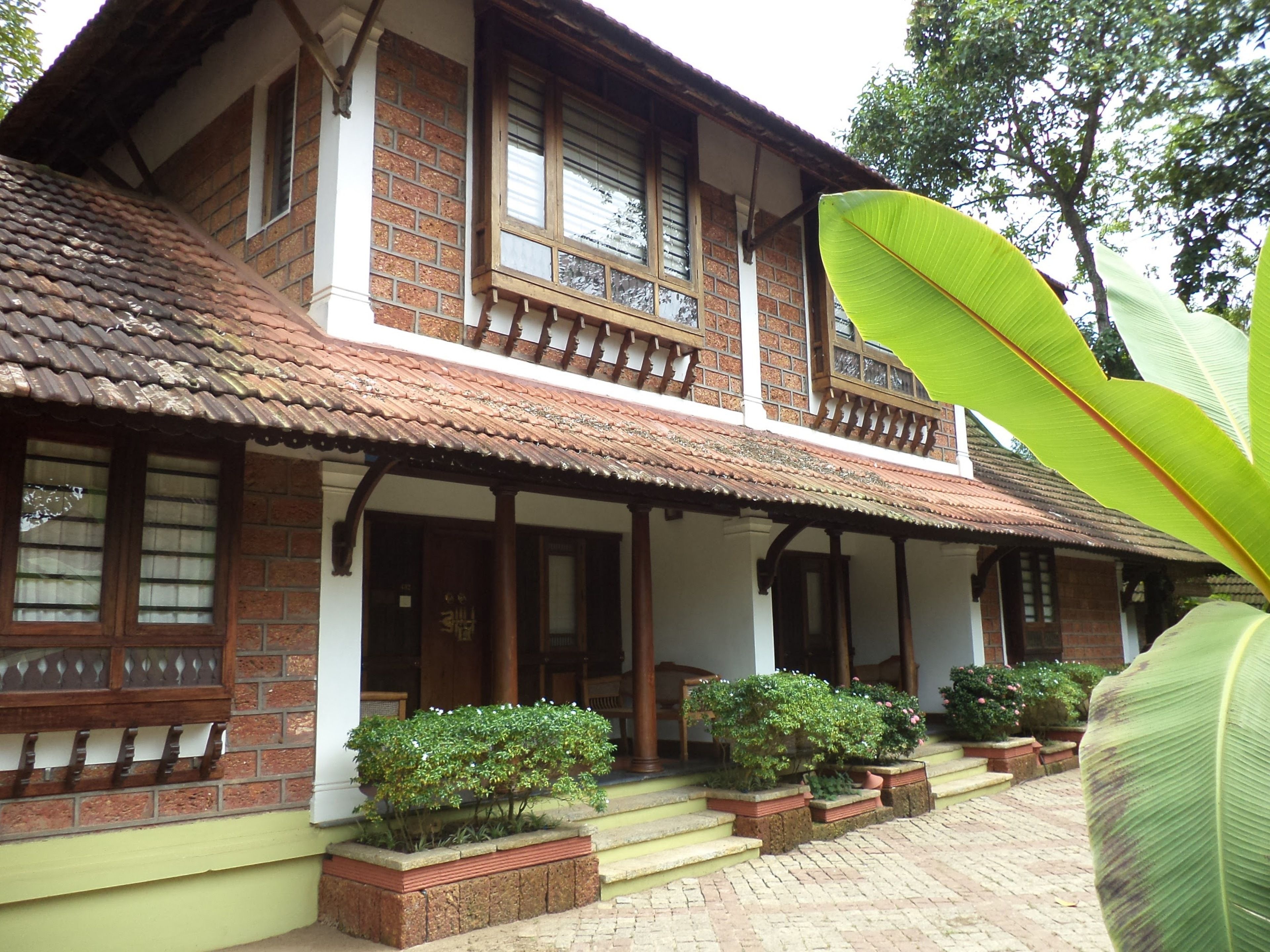 A traditional two-story building with a tiled roof, wooden balconies, and brick walls. There are potted plants and a staircase leading to the entrance.
