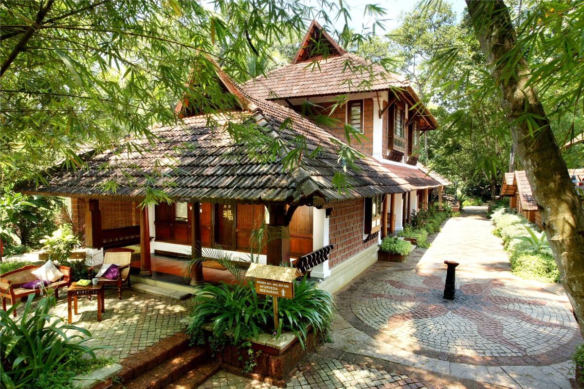 A traditional wooden house with a tiled roof, surrounded by lush greenery and a paved pathway.