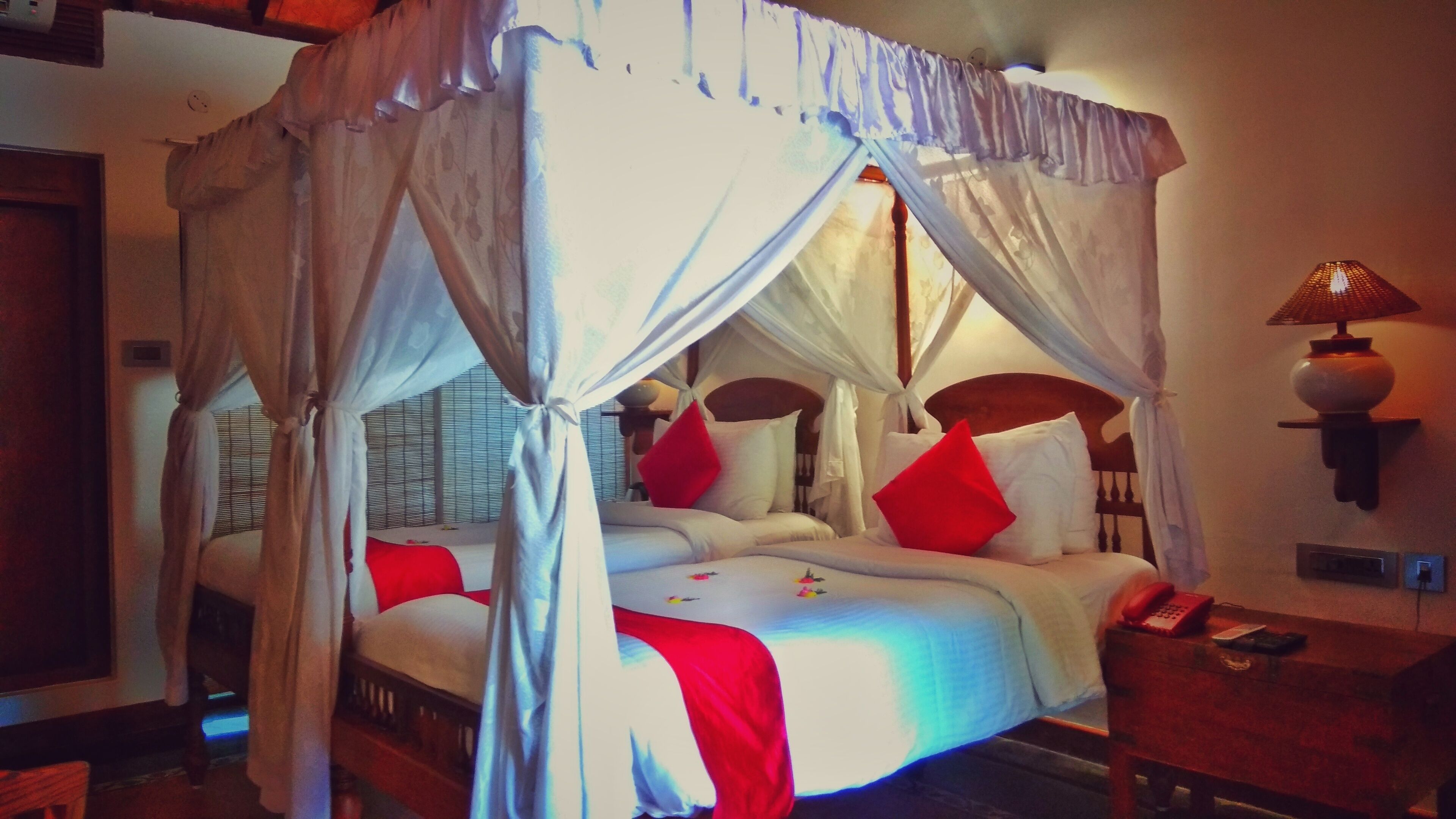 A four-poster canopy bed with white linens and red pillows, a bedside table with a lamp, and a window with blinds.