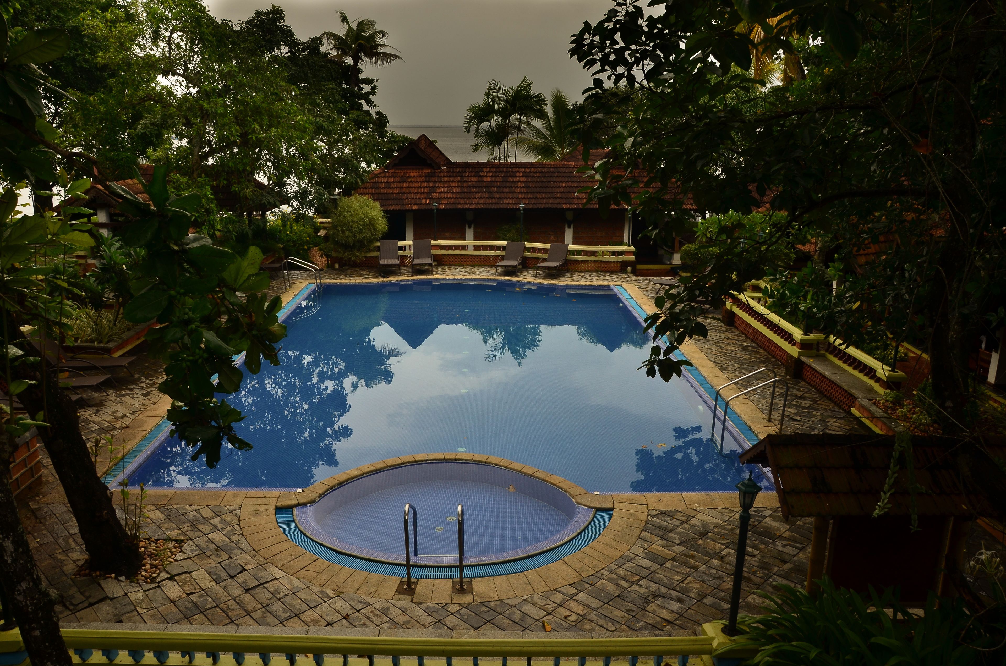 A swimming pool with a tiled deck, surrounded by lush greenery and a thatched-roof building.