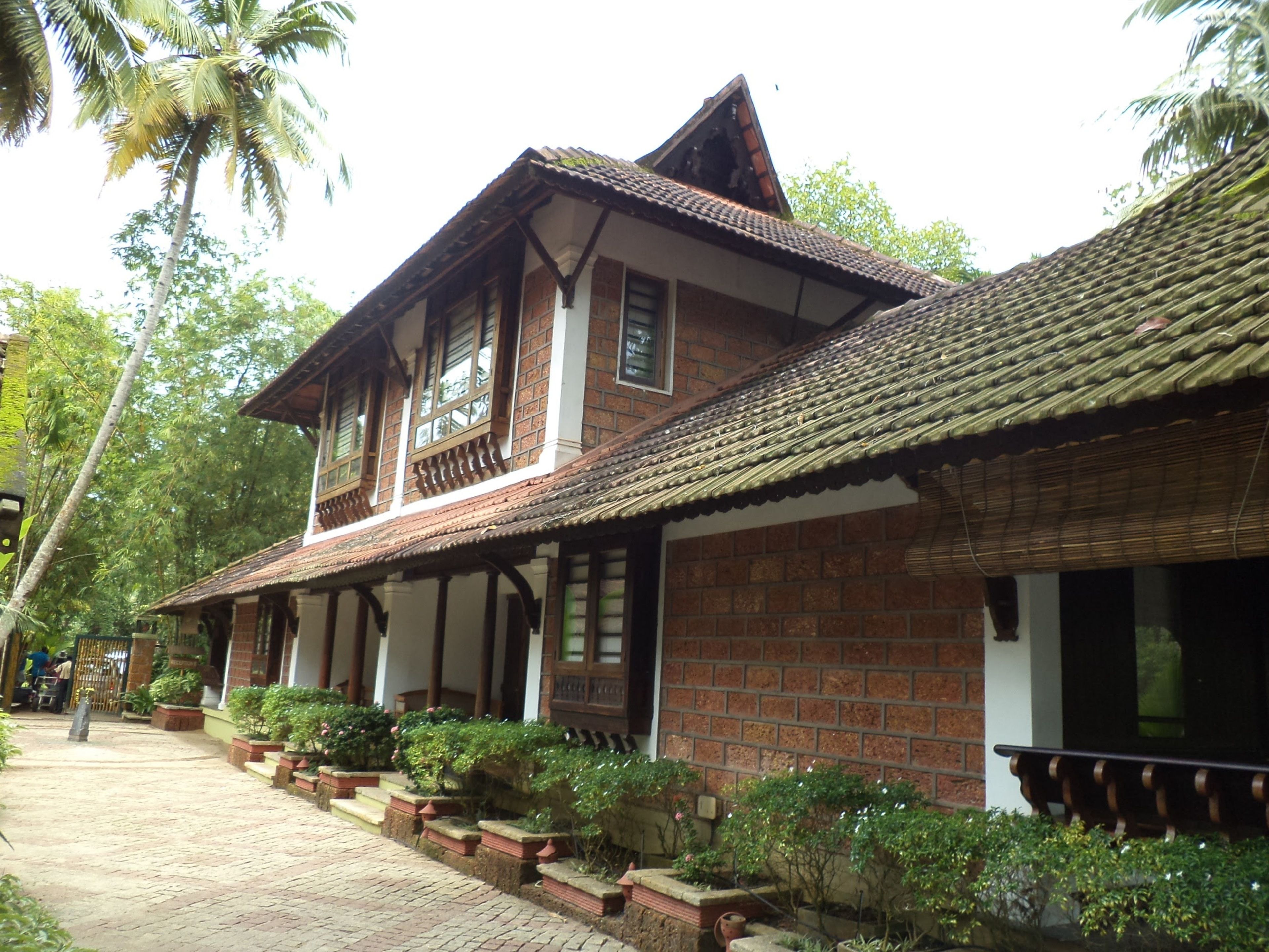 A traditional two-story building with a tiled roof and brick walls, surrounded by lush greenery and palm trees.