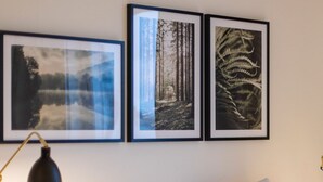 A neatly made bed with white bedding, a black desk lamp, and three framed pictures on the wall above. - Hotell Mörby AB, Patienthotell (Danderyd)