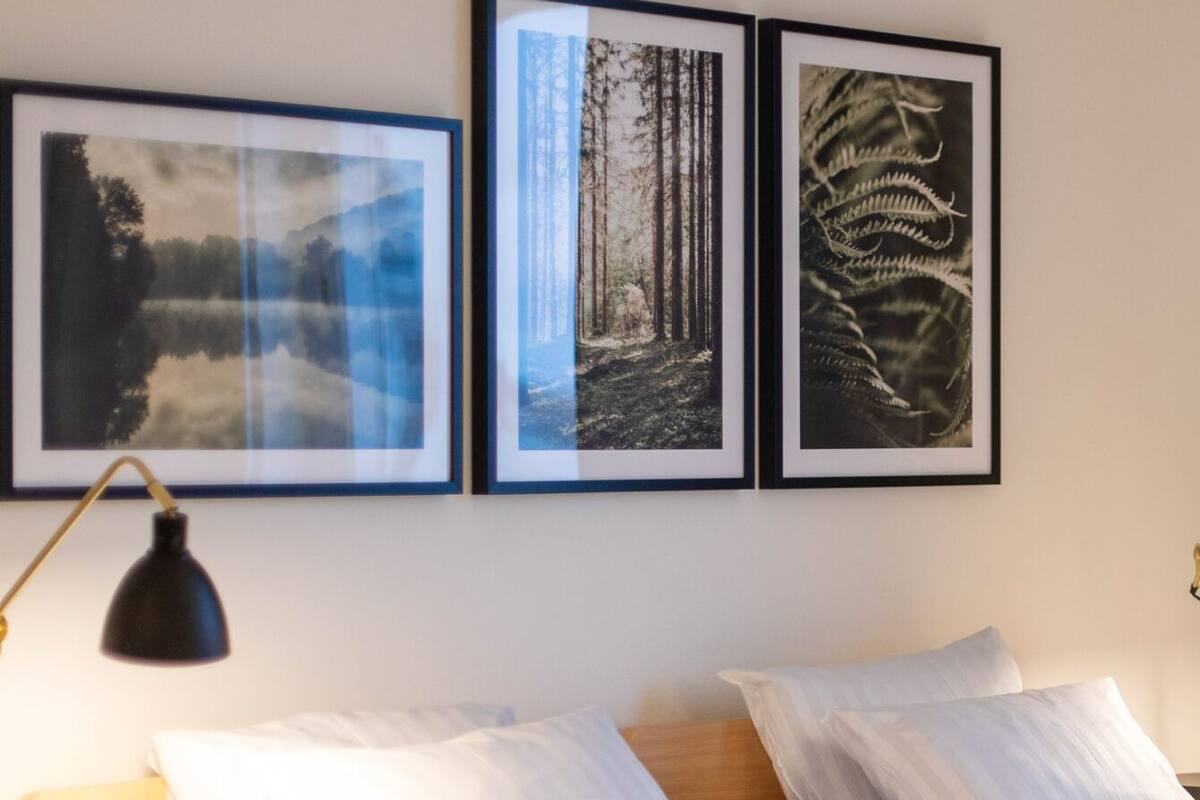 Una cama bien hecha con ropa de cama blanca, una lámpara de escritorio negra y tres cuadros en la pared de arriba.