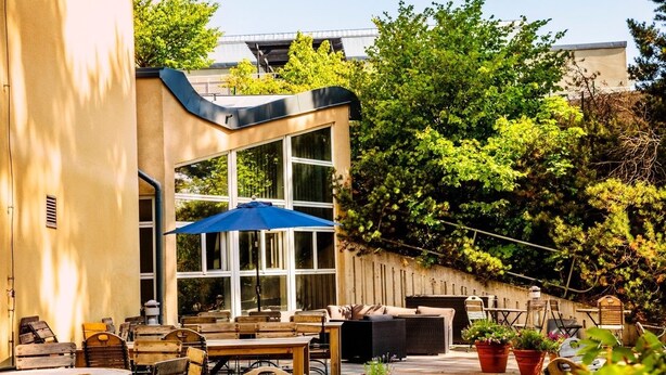 An outdoor patio area with wooden furniture, a blue umbrella, and potted plants. - Hotell Mörby AB, Patienthotell (Danderyd)